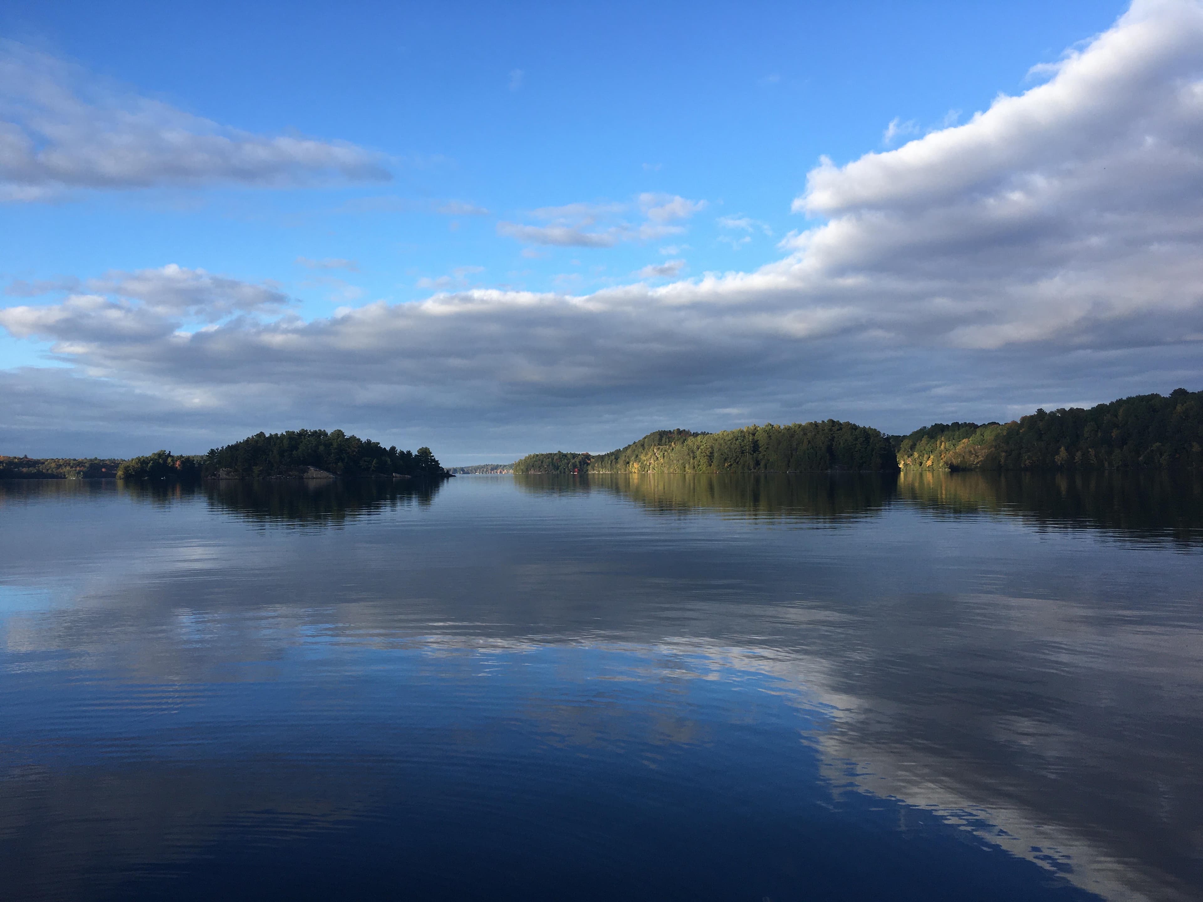 Serene lake with forested islands reflecting a blue sky filled with soft white clouds.