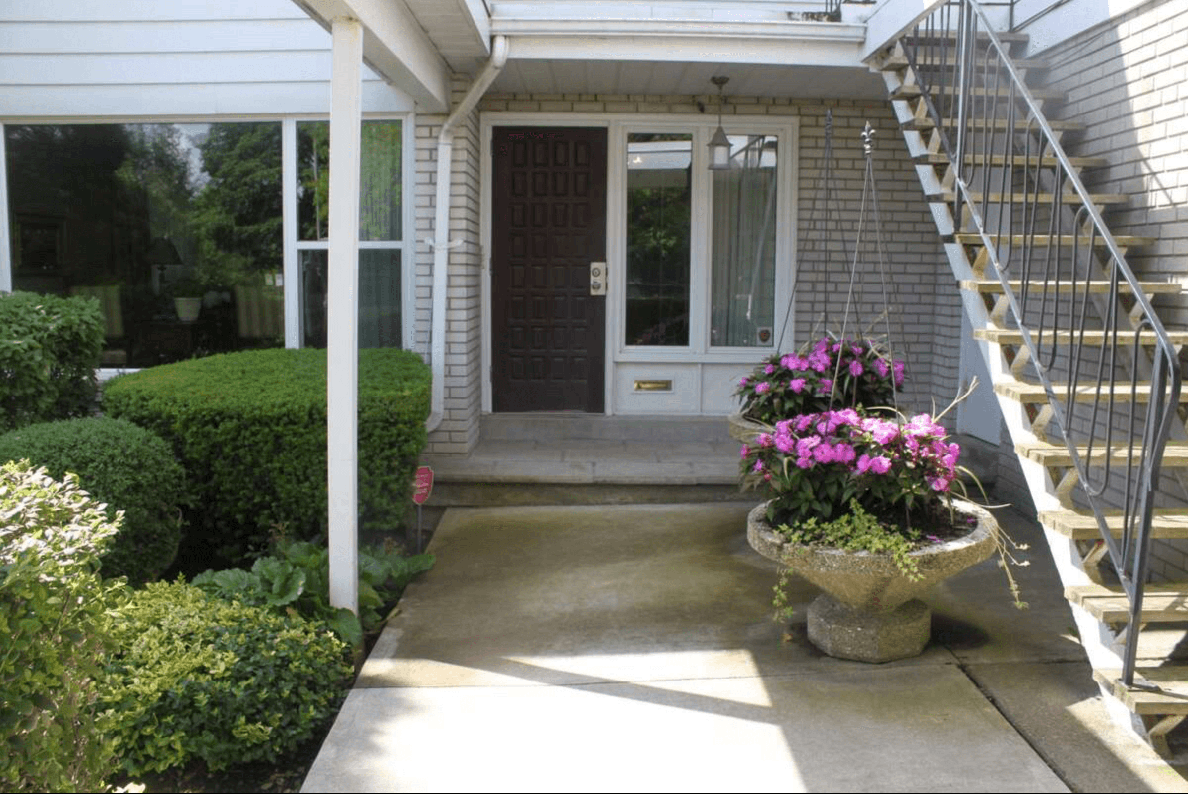 Concrete walkway leading to a brown front door flanked by green bushes and pink flowers.