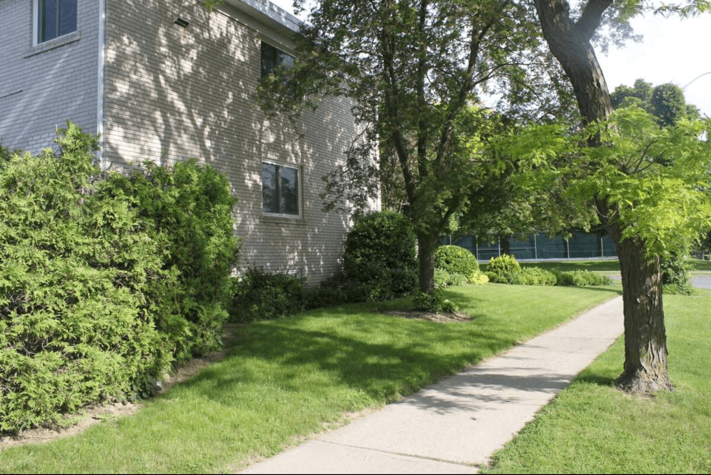 Concrete sidewalk alongside a light brick building with lush green grass and leafy trees.