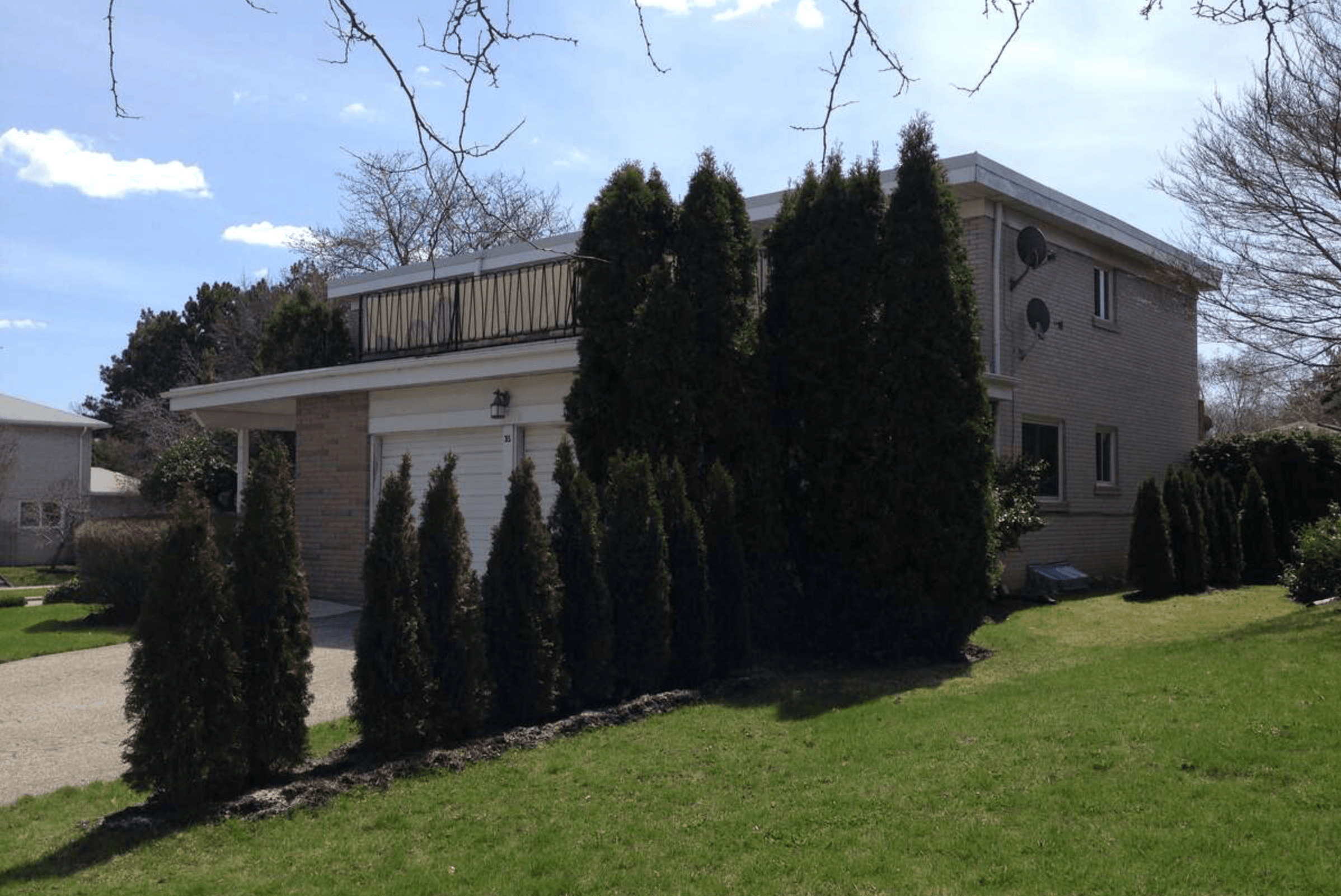 Two-story house with a flat roof balcony and tall evergreen trees lining the yard.