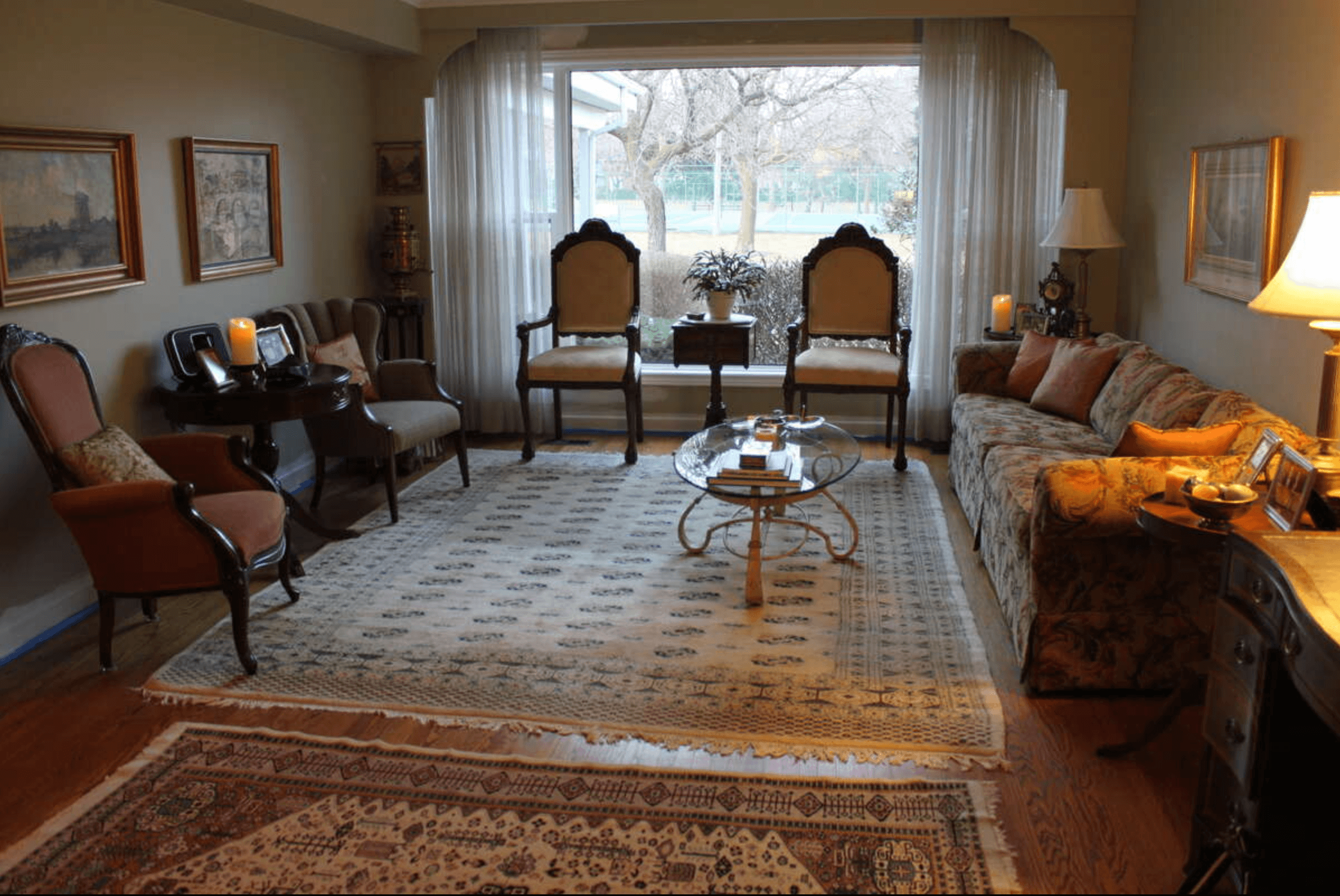 Traditional living room with floral sofa, patterned rugs, and large window overlooking a backyard.