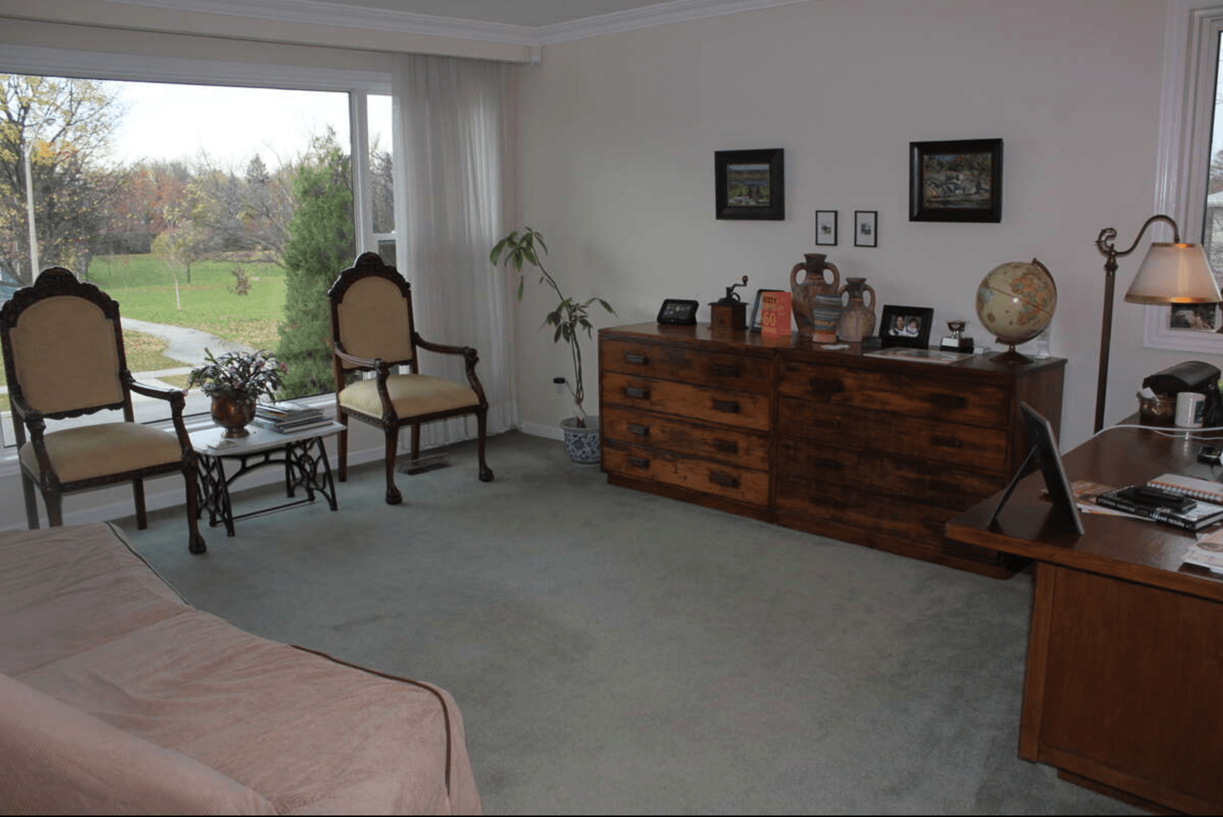 Sunlit room with two ornate armchairs by a large window, a wooden dresser, and a desk.