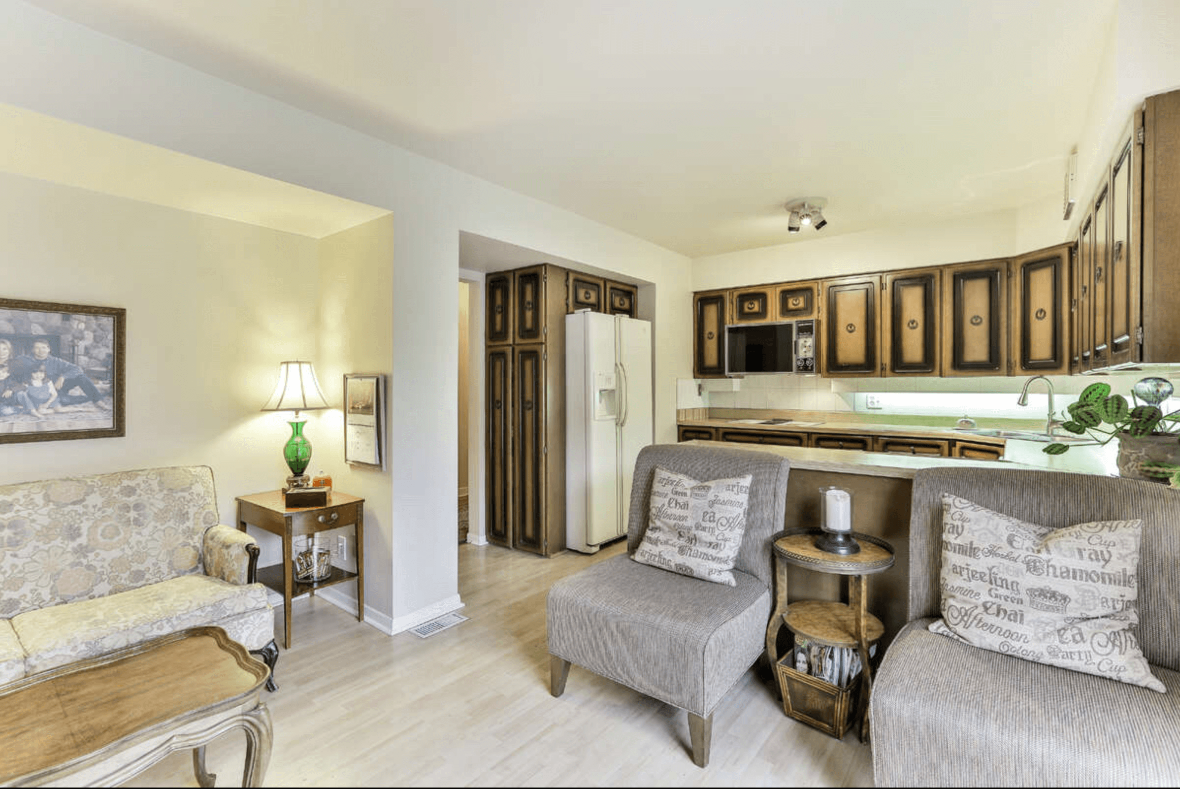 Living room with floral sofa and grey armchairs next to kitchen with dark wood cabinets.