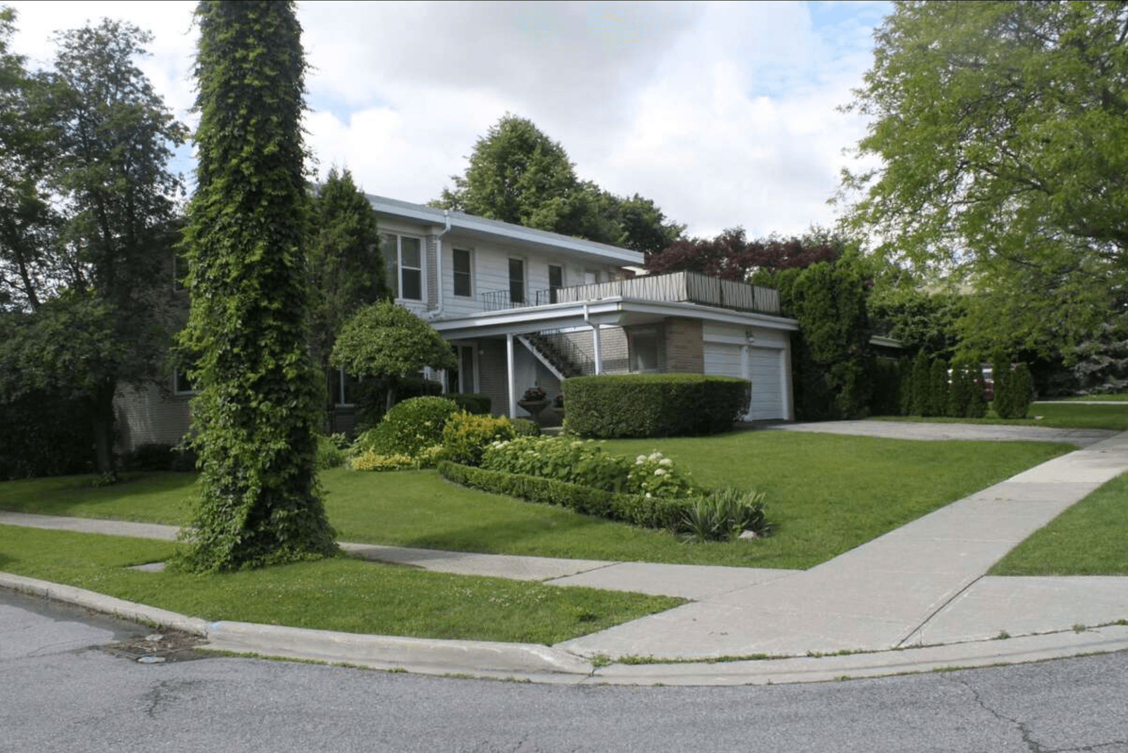 Two-story suburban house with a manicured lawn, lush landscaping, and a tall vine-covered tree.