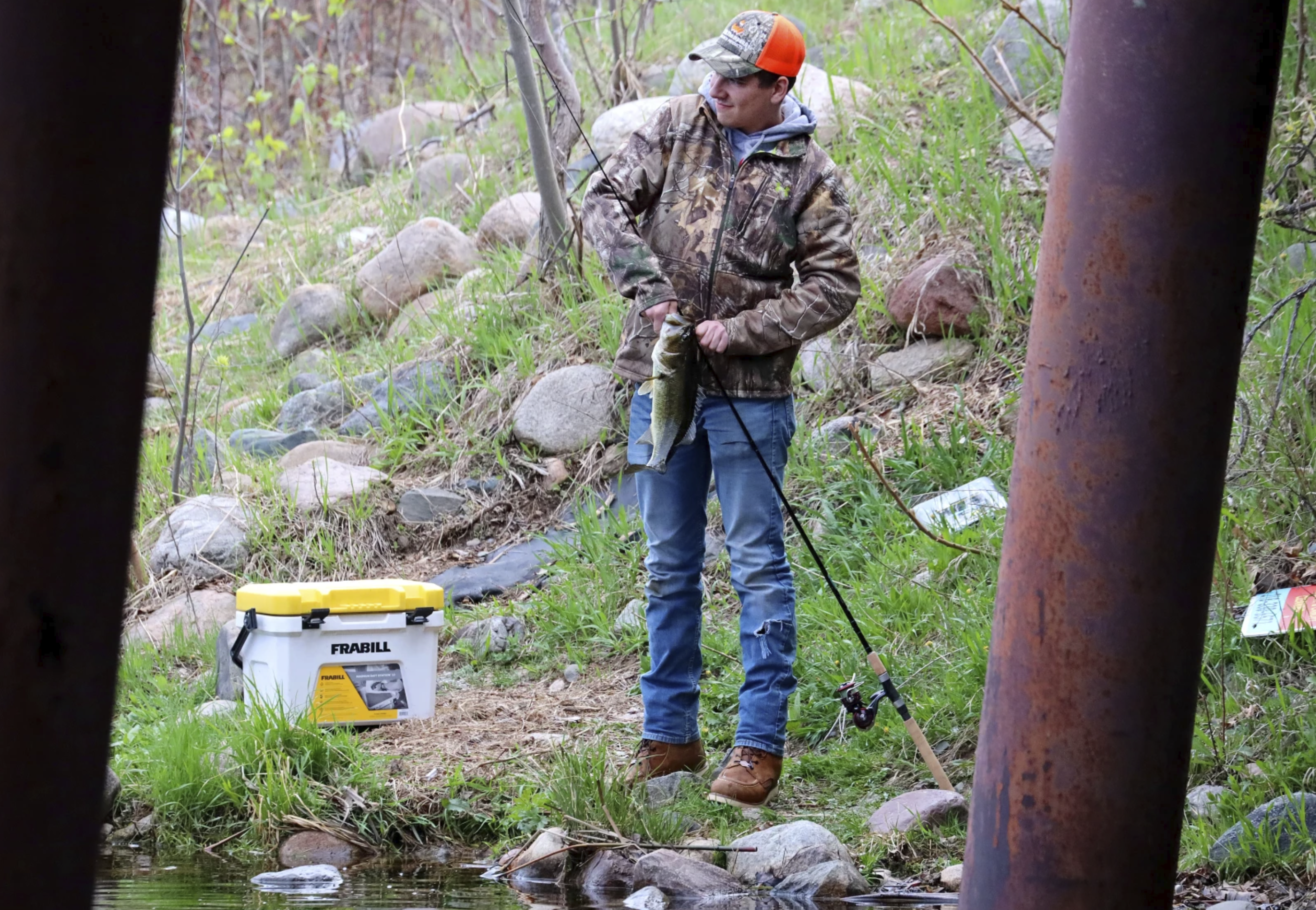 Fisherman in camouflage holds a caught bass on a rocky riverbank near a white cooler.