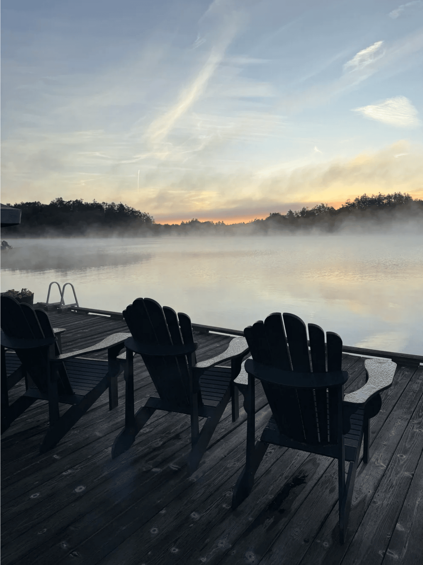 Three Adirondack chairs on a wooden dock overlook a misty lake at sunrise.