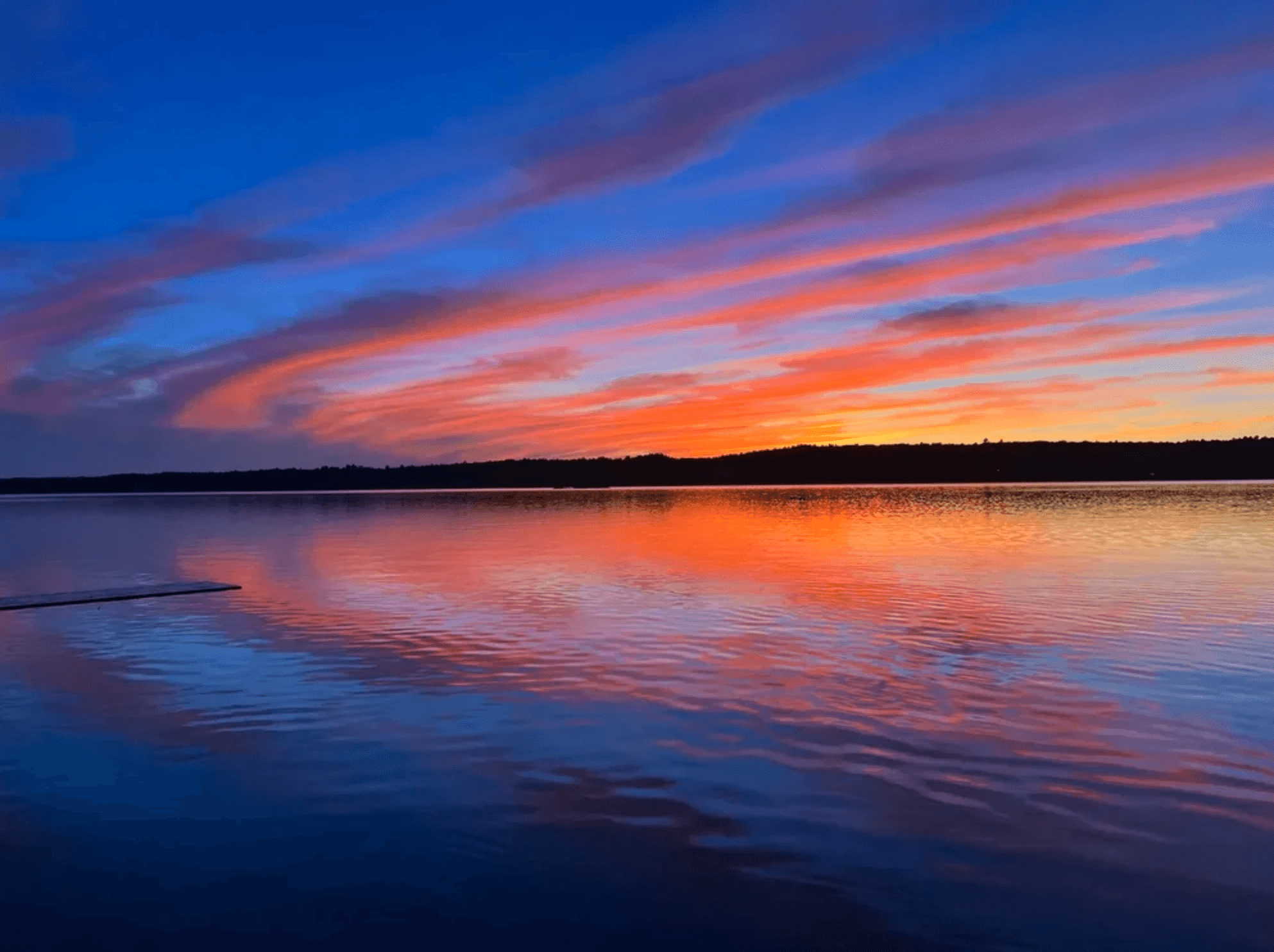 Vibrant orange and pink sunset clouds reflecting on a calm lake with a dark shoreline.