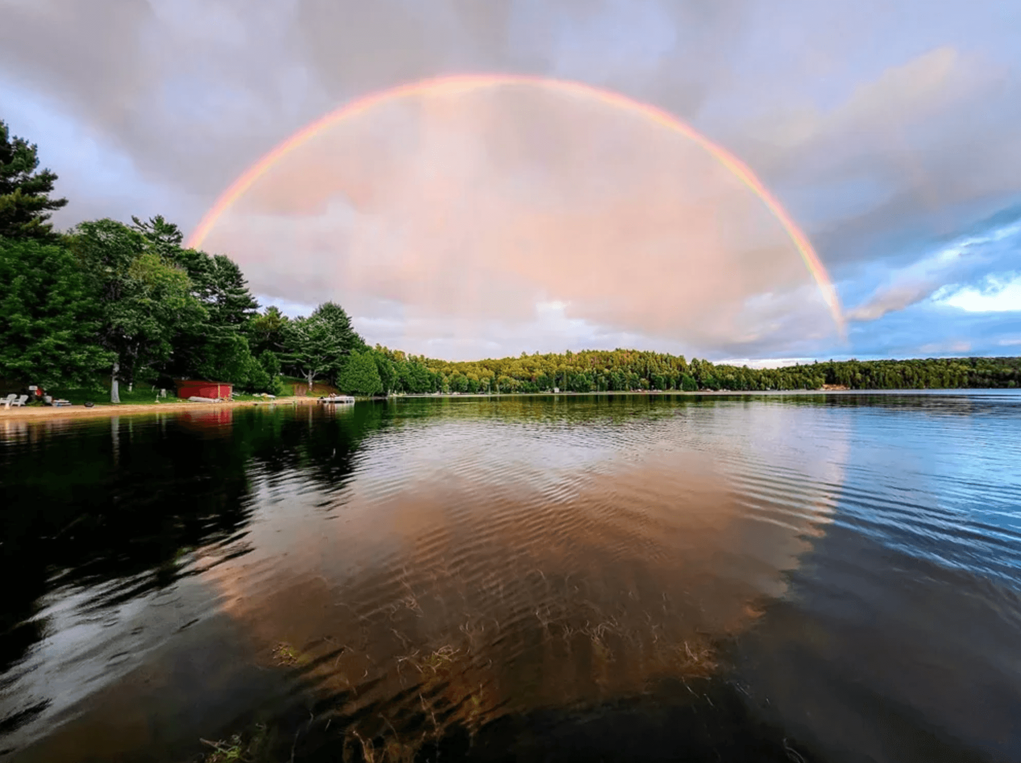 Vibrant rainbow arcs over a calm lake, reflecting in the water near a forested shoreline.