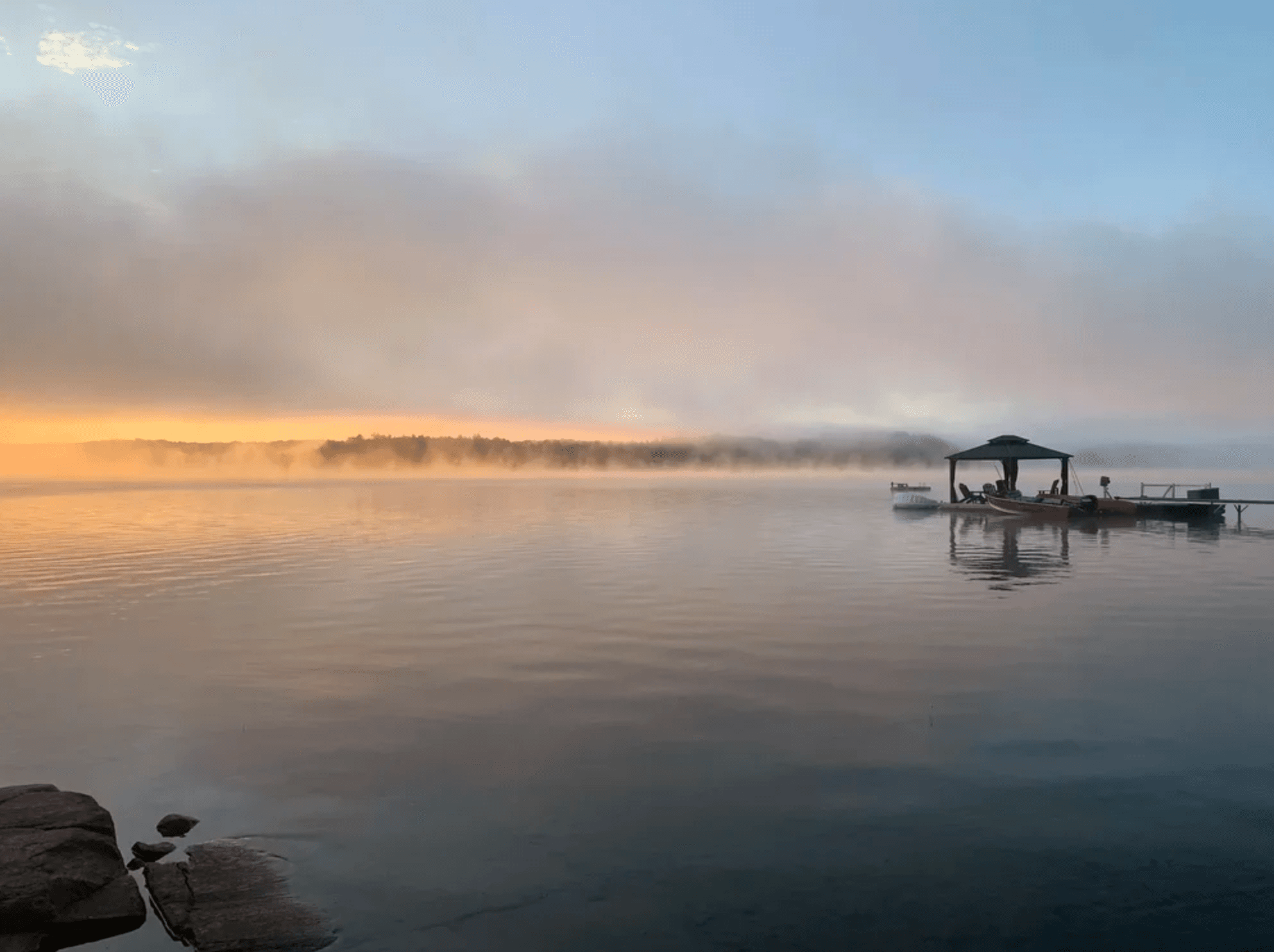 Misty sunrise over a calm lake with a gazebo on a dock and golden horizon.