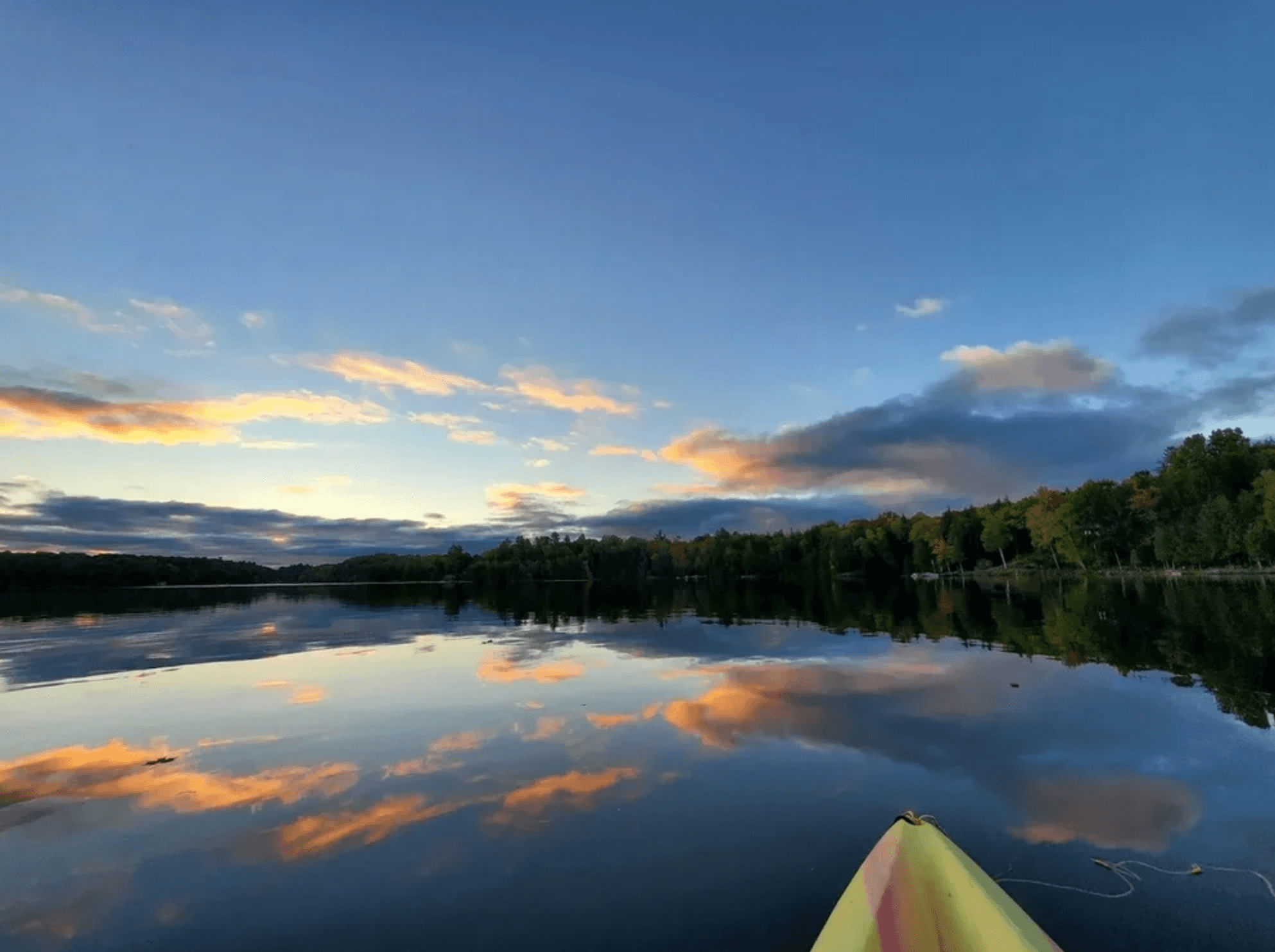 Sunset clouds reflect on a calm lake, viewed from the bow of a yellow kayak.
