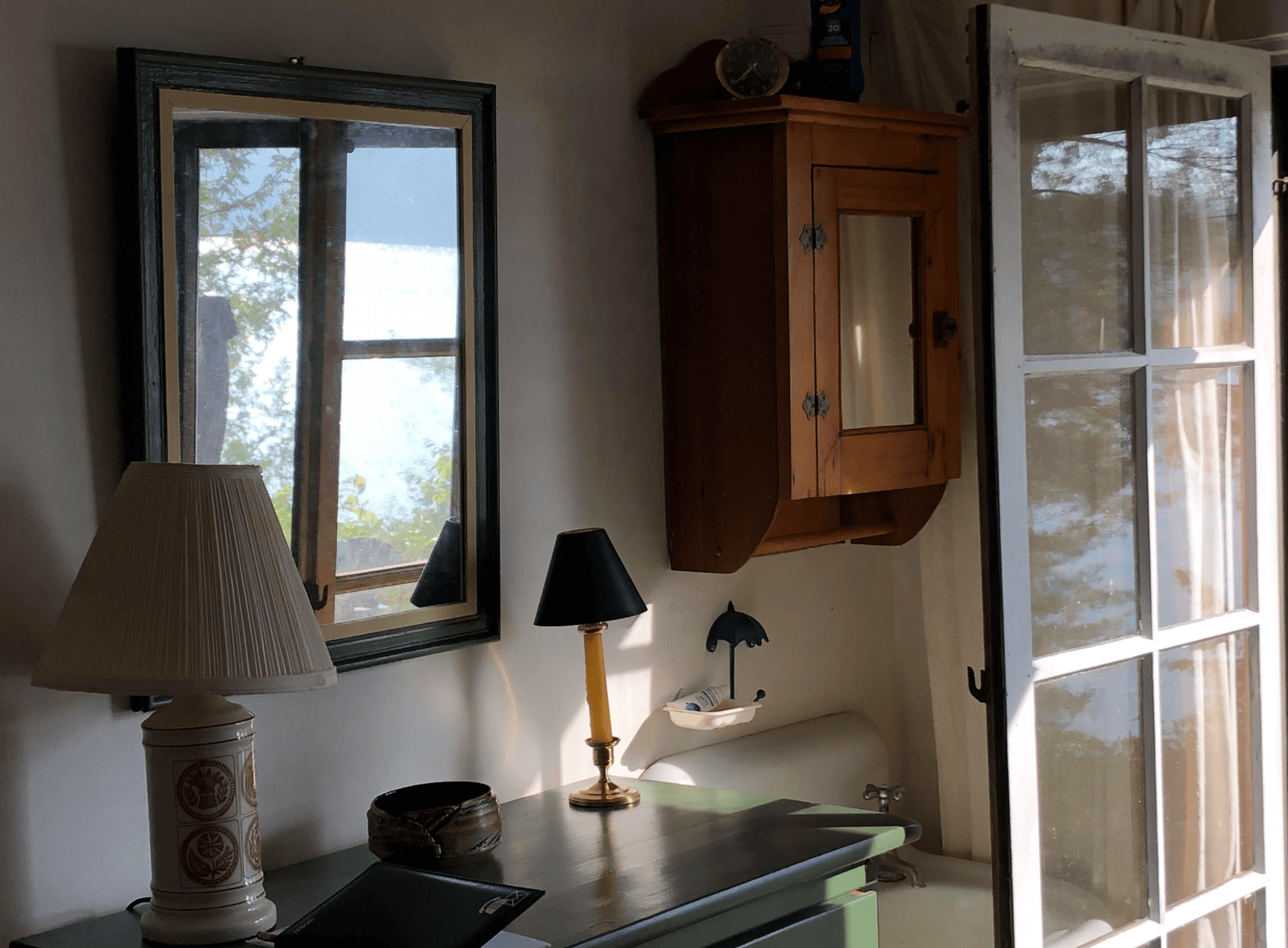 Sunlit rustic interior with a mirror reflecting a lake, two lamps, and a wooden cabinet.