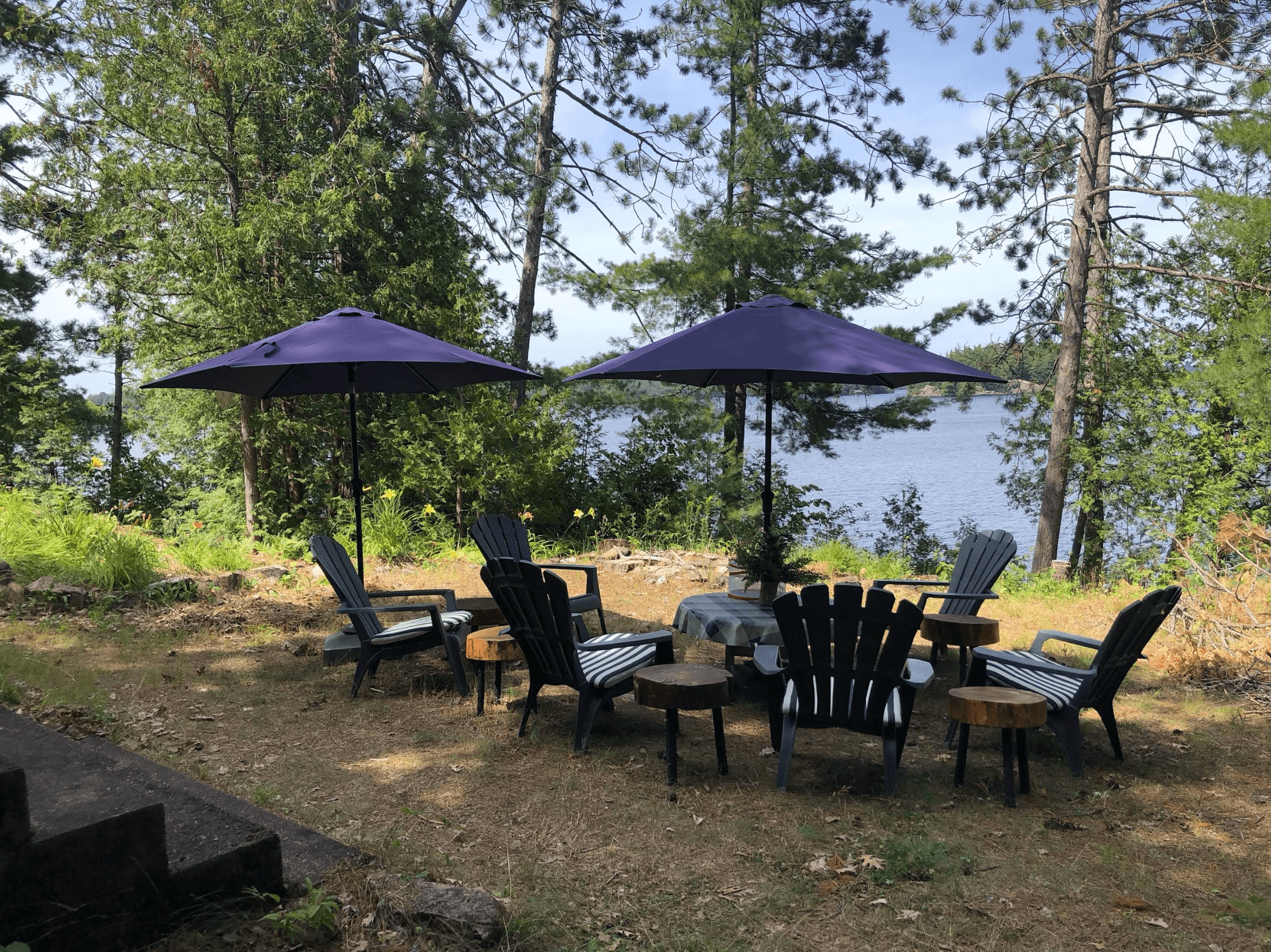 Two purple umbrellas shade Adirondack chairs and small tables on a wooded lakeside patio.