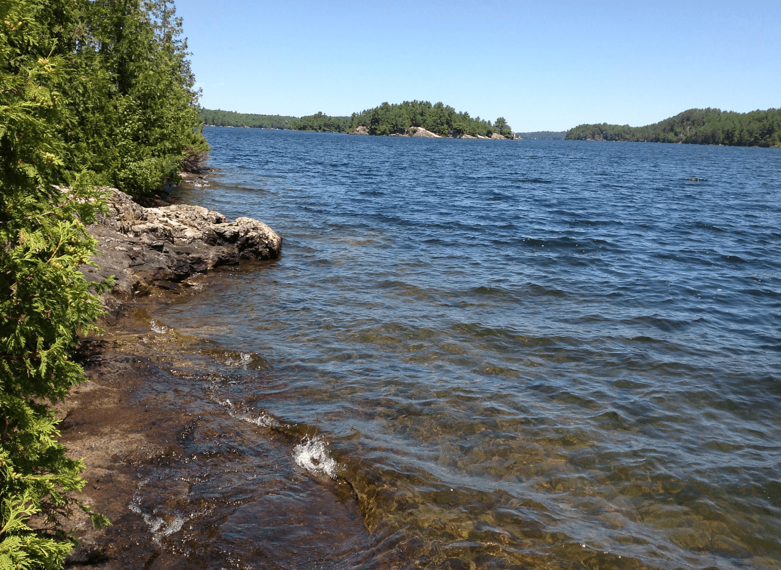 Rocky lake shoreline with clear water, green trees, and distant forested islands under blue skies.