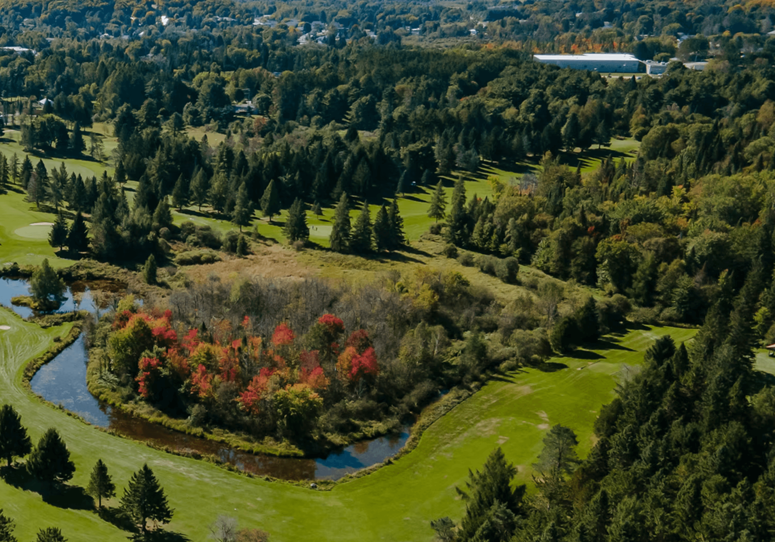 Aerial view of a green golf course with a winding stream and colorful autumn trees.