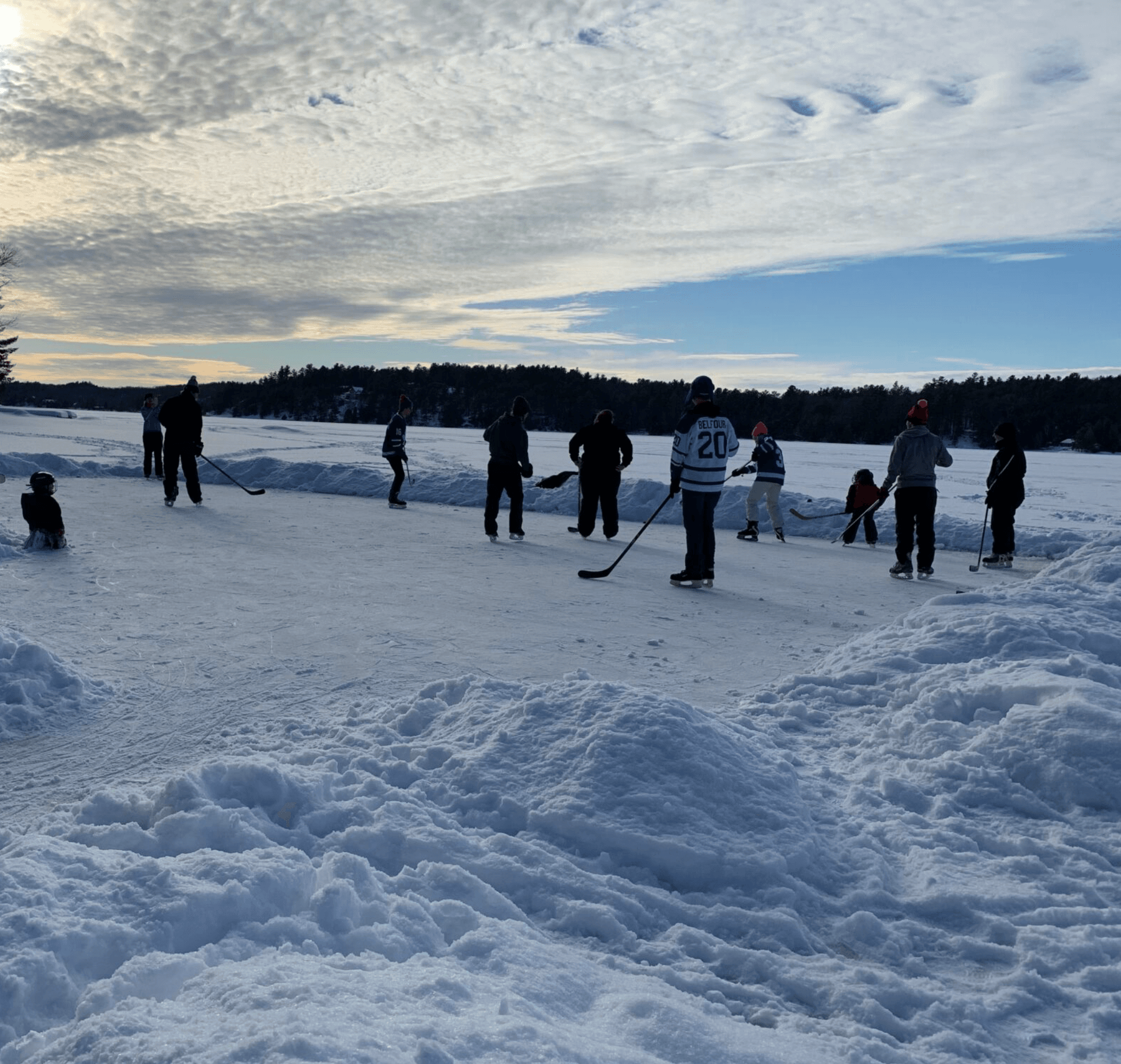 People playing ice hockey on a frozen lake rink surrounded by snow and trees.