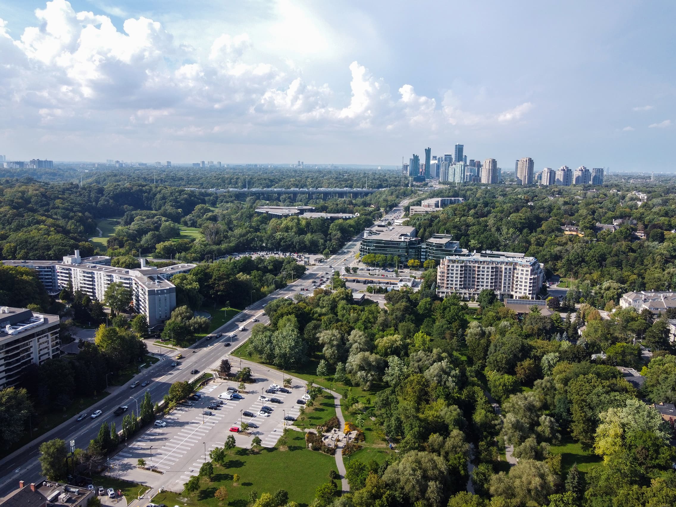 Aerial view of a lush green city with roads, buildings, and a distant skyscraper skyline.