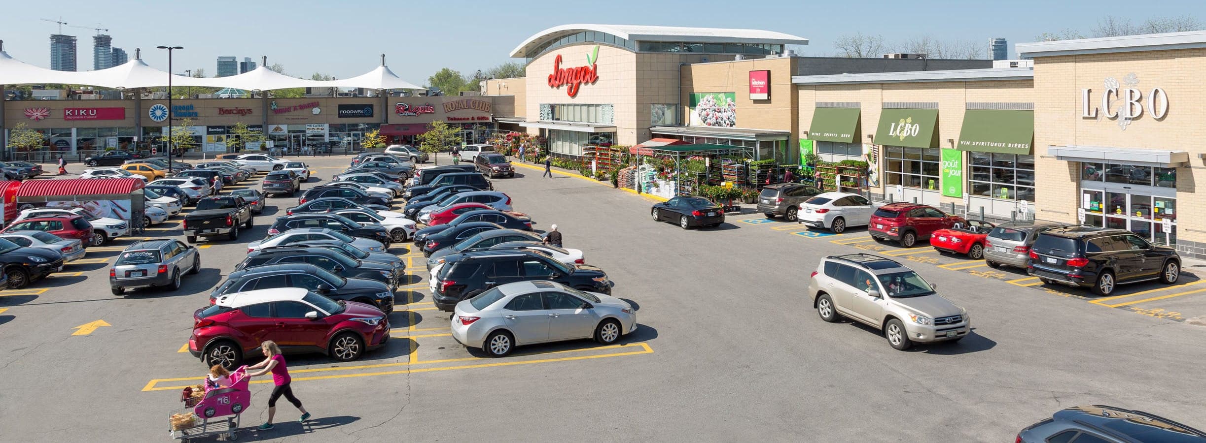 Busy shopping plaza parking lot with Longo's and LCBO storefronts on a clear sunny day.